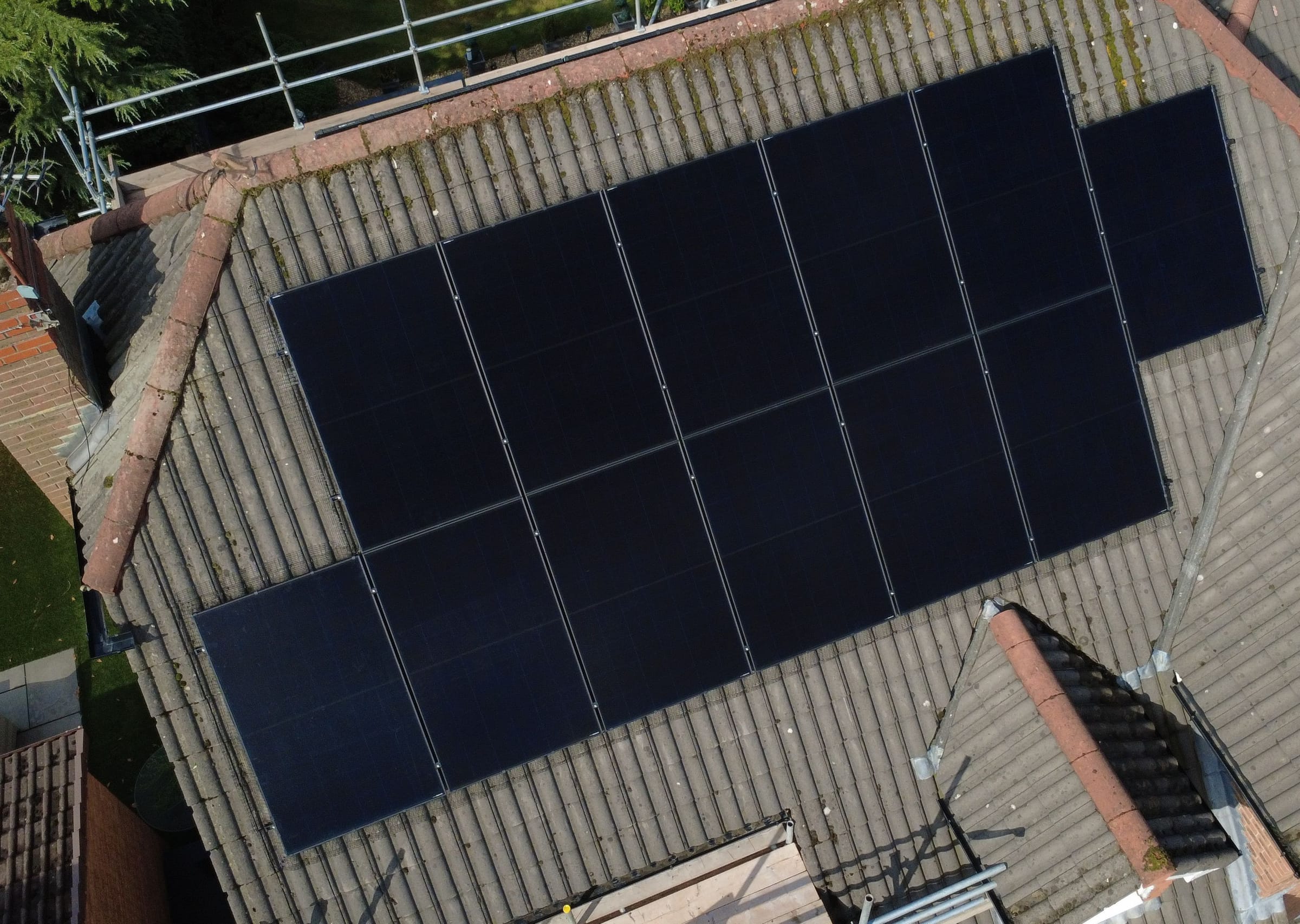 A long array of black panels across a grey tiled roof, seen from a drone above the garden